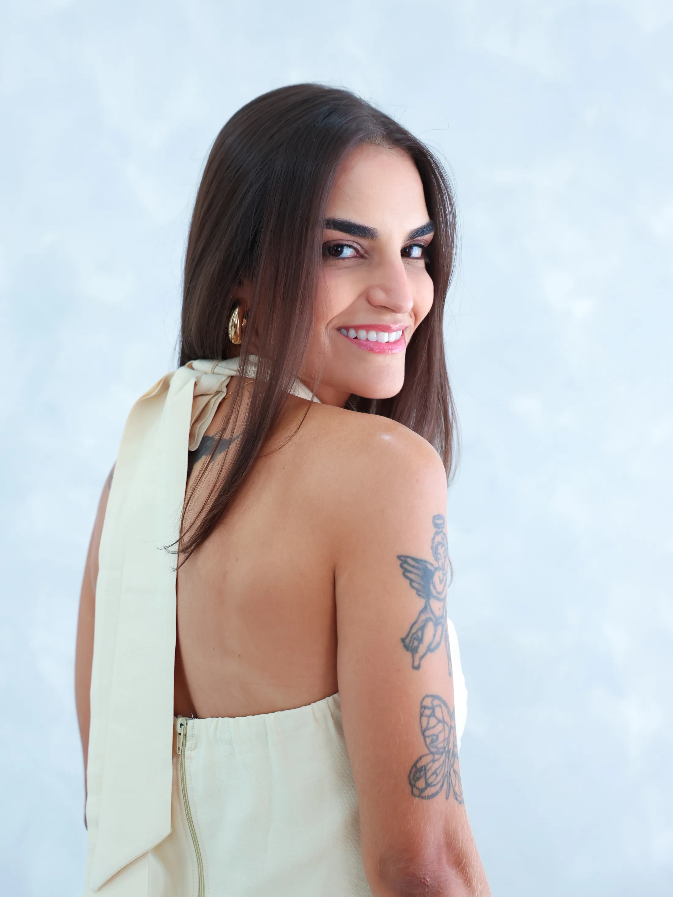 Mulher de roupa clara sorri por cima do ombro, diante de fundo azul acinzentado.