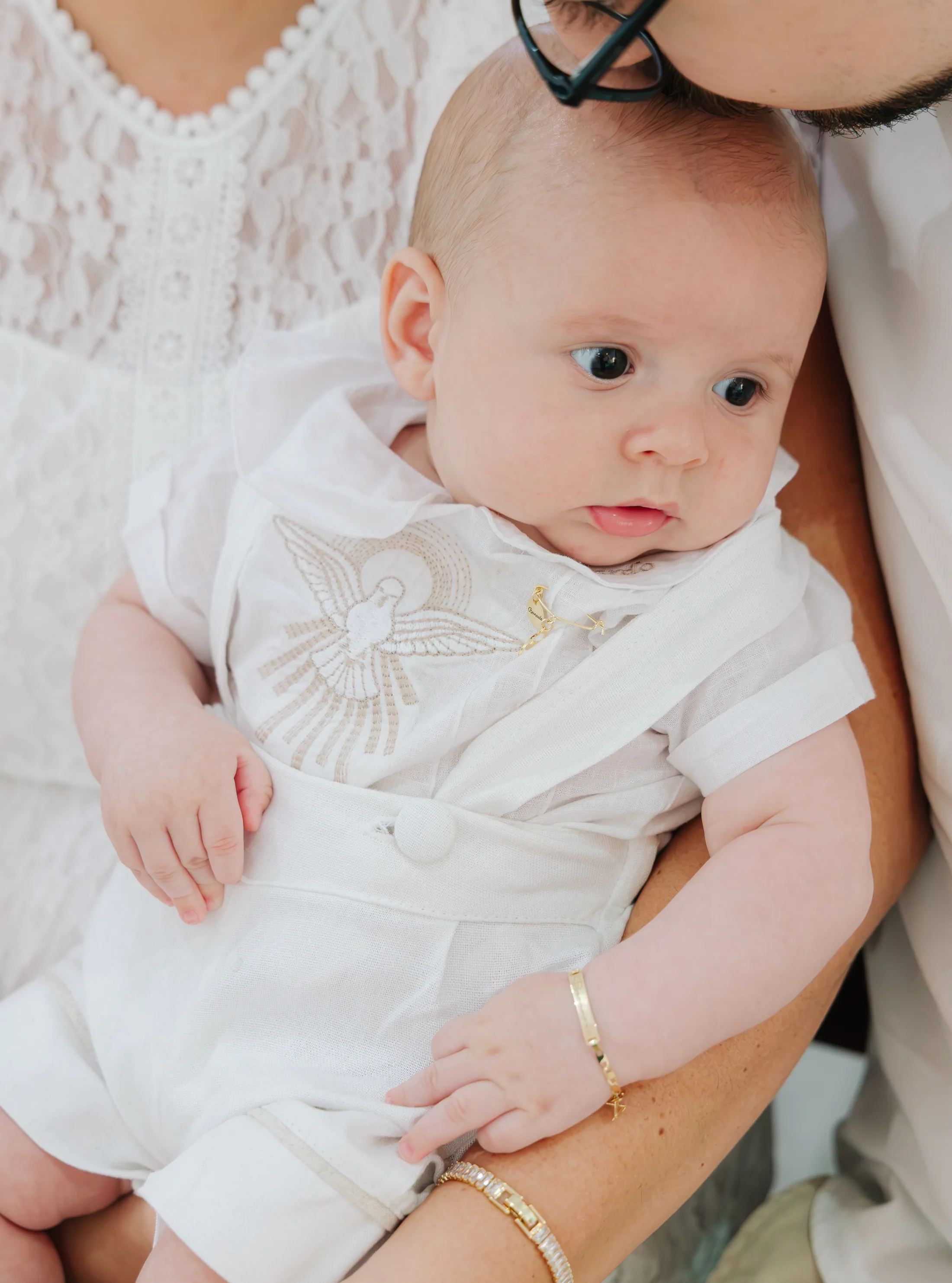 Bebê de roupa branca está no colo de uma pessoa com vestido rendado.