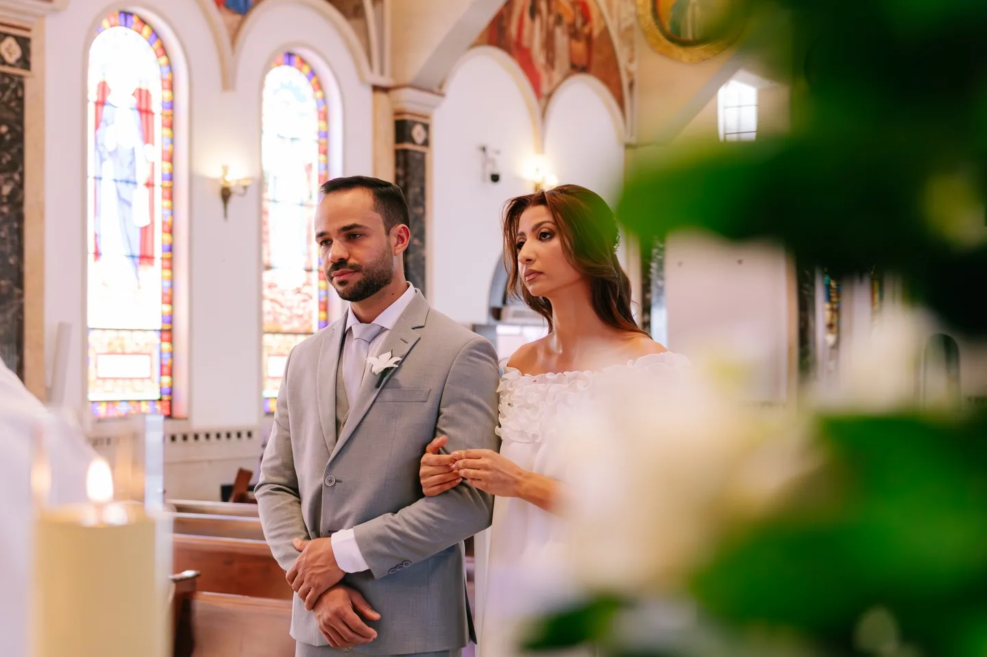 Casal de braços dados em uma igreja, visto entre flores desfocadas no primeiro plano.