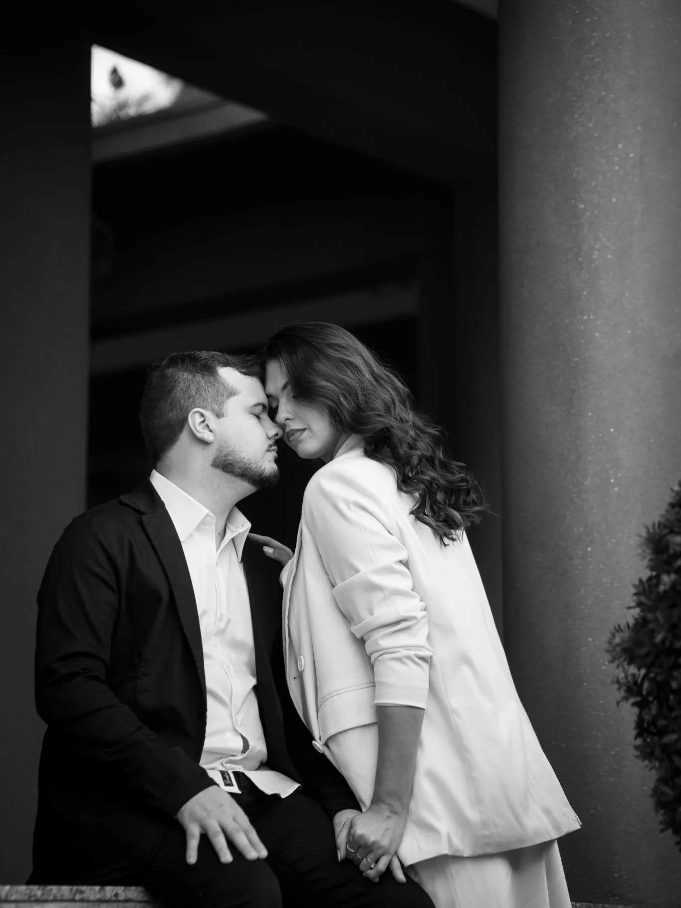 Casal aproxima os rostos em uma entrada sombreada, em fotografia preta e branca.