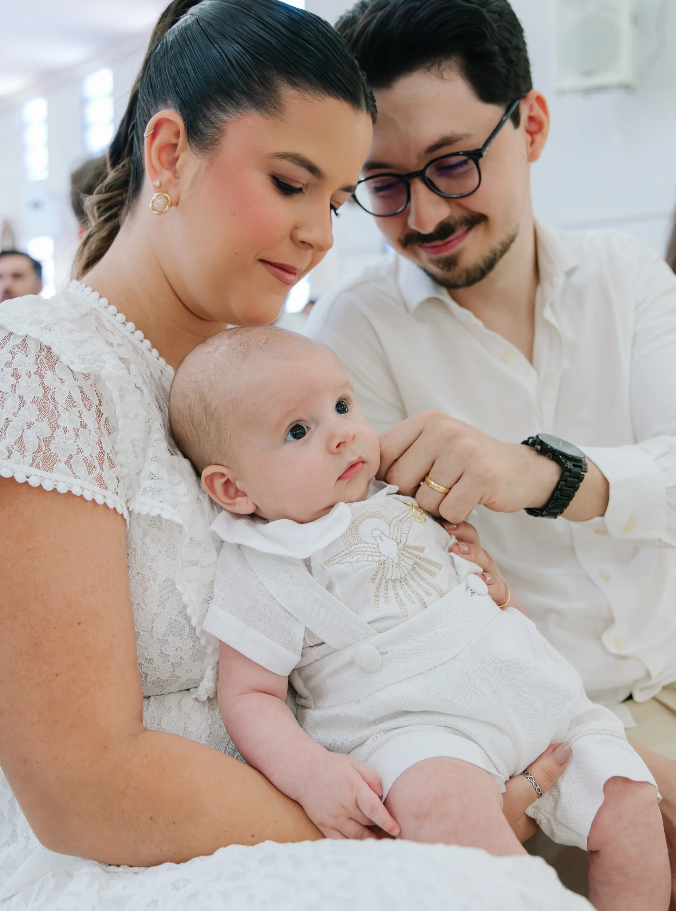 Mulher e homem olham para um bebê vestido de branco no colo da mulher.
