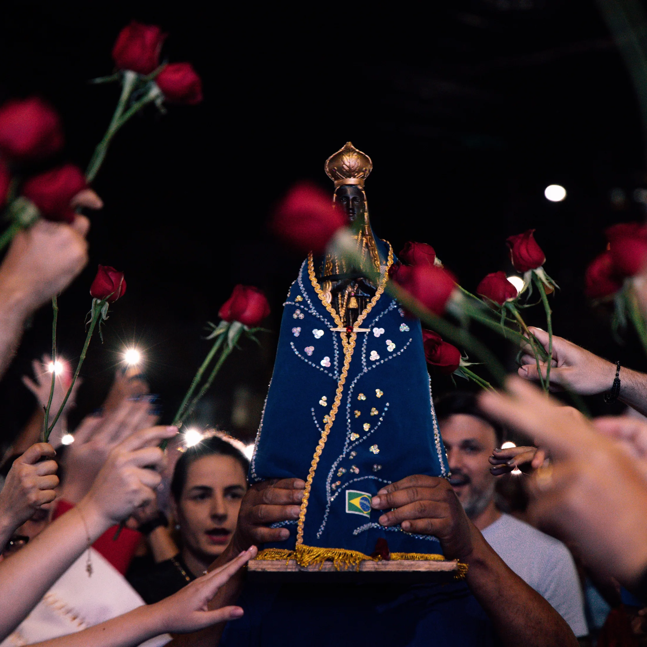 Imagem religiosa de manto azul é carregada entre pessoas que erguem rosas vermelhas.