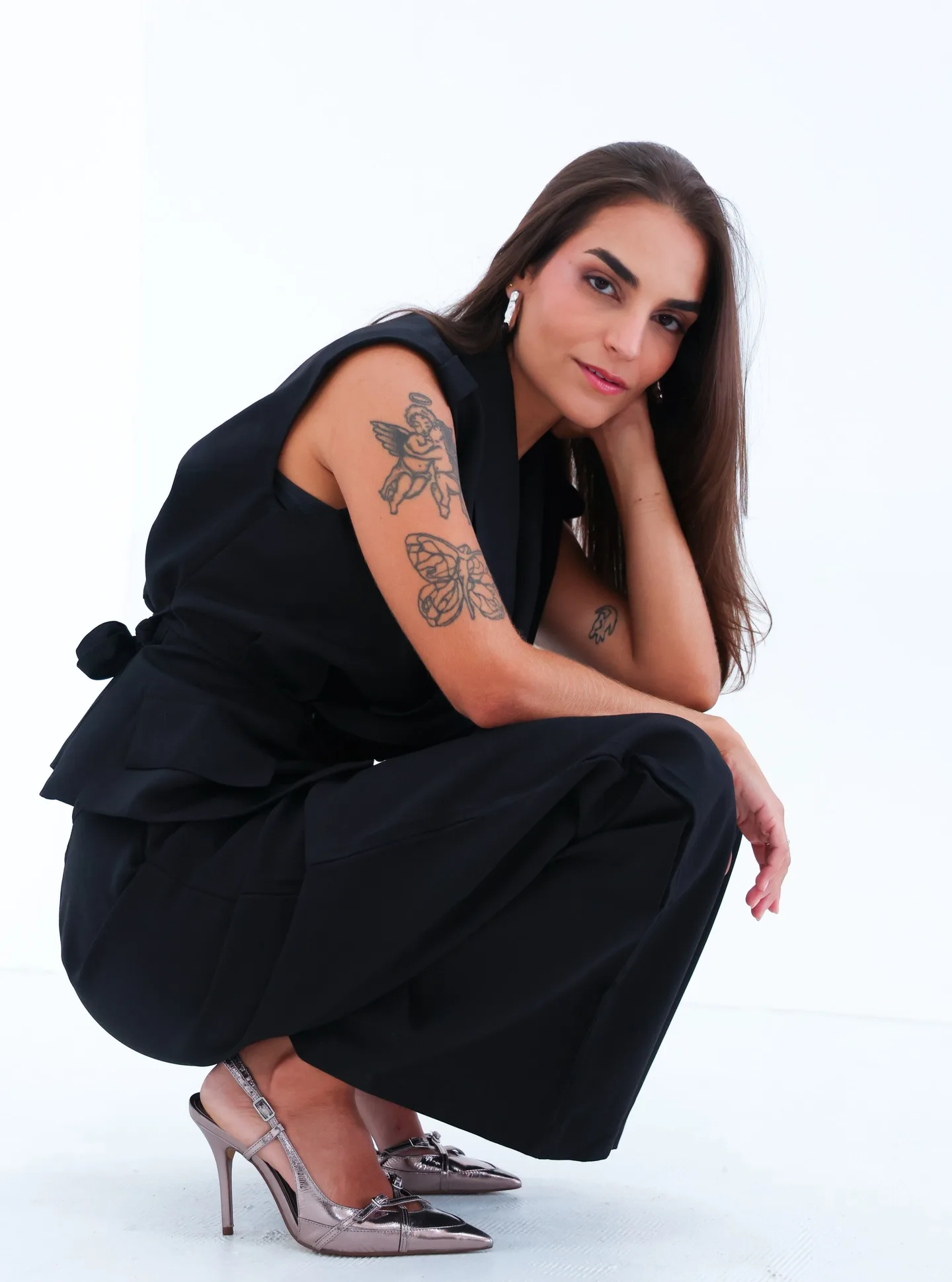 Mulher de roupa preta posa agachada diante de fundo branco.