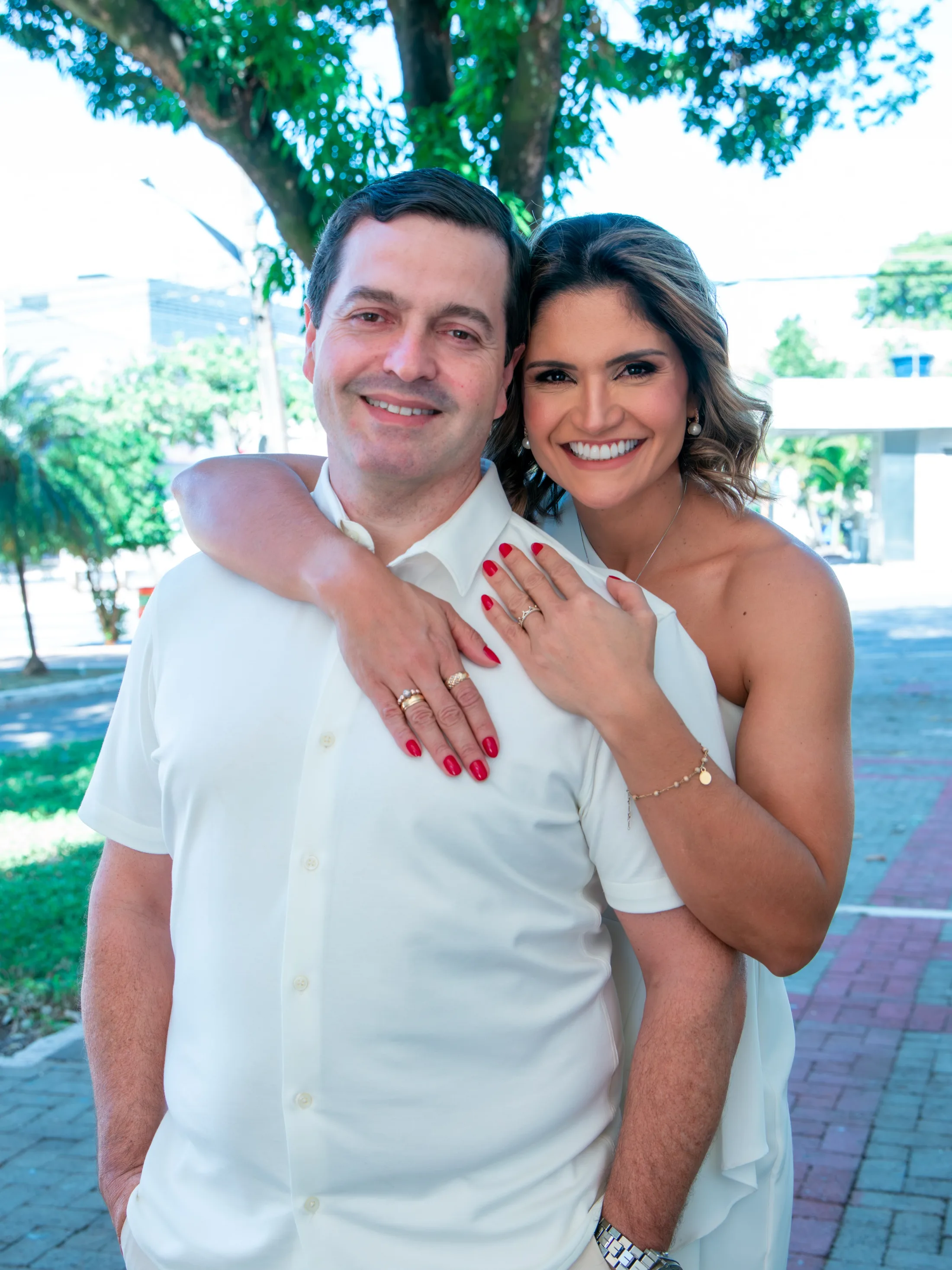 Casal de roupa branca sorri abraçado sob a sombra de uma árvore.