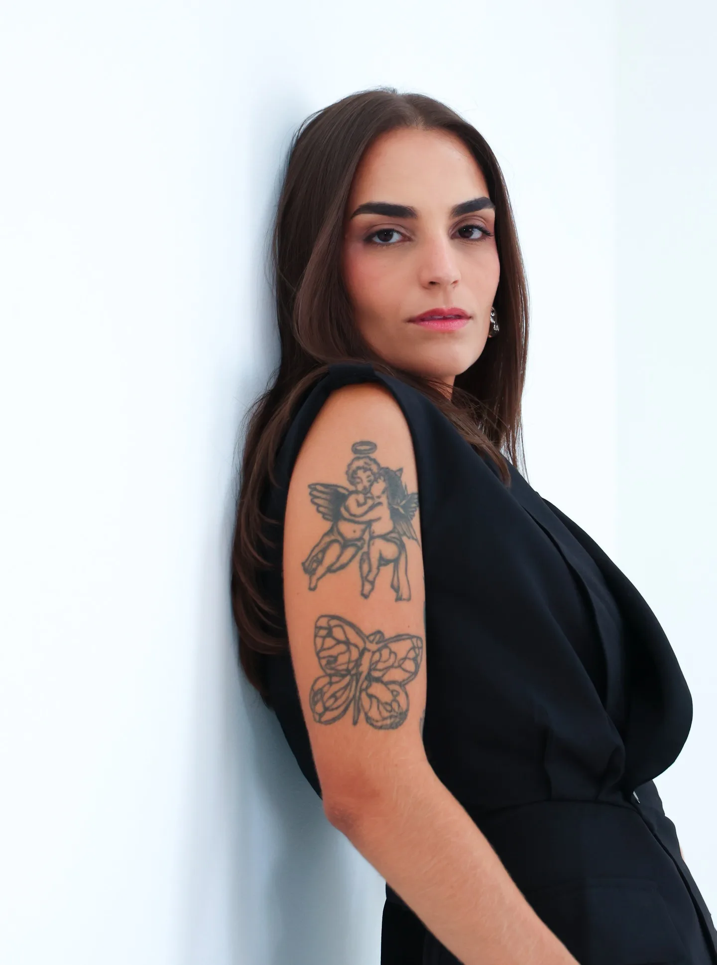 Mulher de roupa preta posa de perfil diante de fundo claro, com tatuagens visíveis no braço.