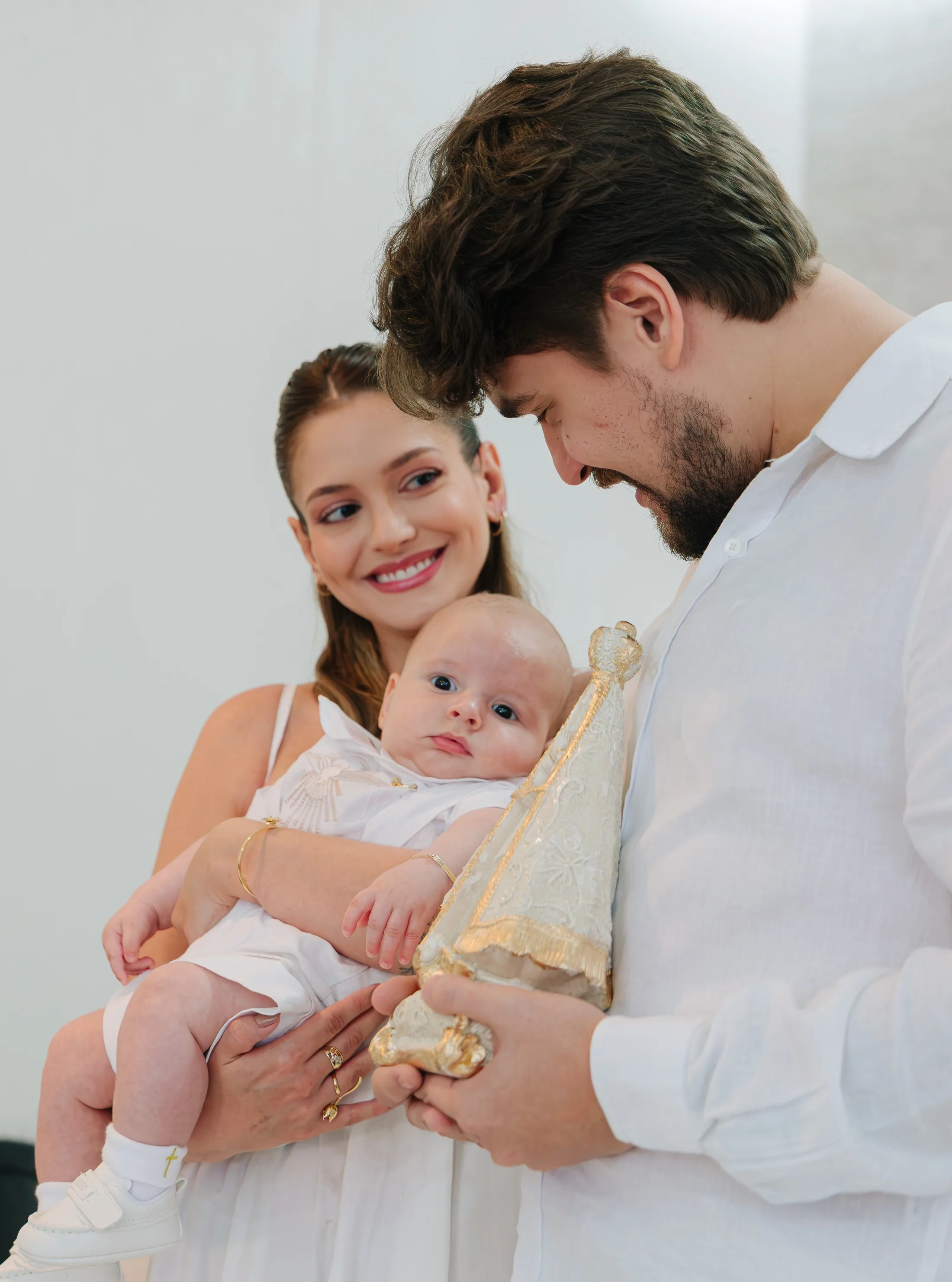 Mulher e homem seguram um bebê diante de uma parede clara.