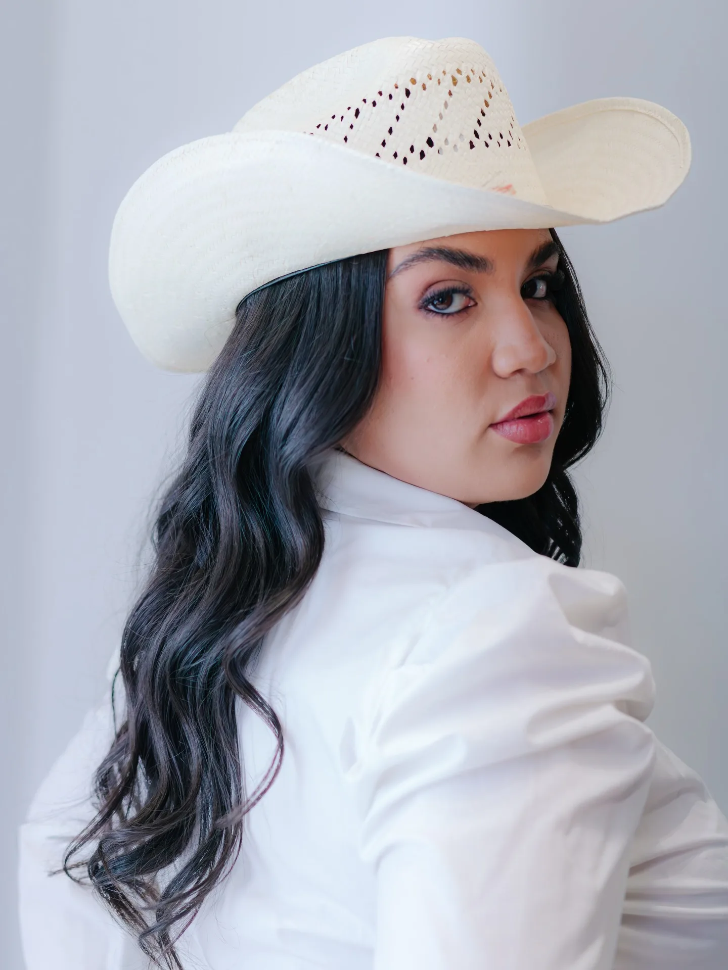 Mulher de chapéu branco olha por cima do ombro diante de fundo cinza.